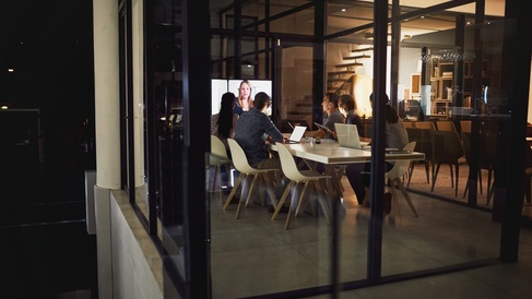 People in a hybrid meeting.  They are sitting round a table looking at a screen at the end of the table showing a remote attendee..